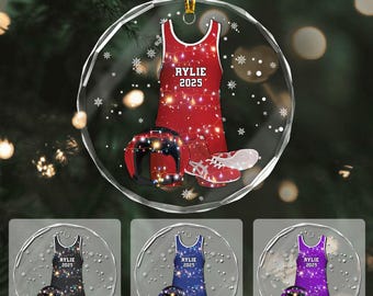Puede incluir: Adorno navideño redondo y transparente con un maillot de lucha libre rojo, un casco y zapatos. El maillot lleva el nombre "RYLIE" y el año "2025". Se muestran adornos adicionales en negro, azul y morado debajo.