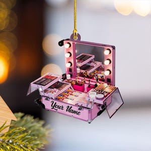 May include: Pink makeup case ornament with a mirror and lights. The case is open, revealing various makeup products. The text "Your Name" is printed on the front of the case. A gold string is attached for hanging.