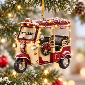 A decorative ornament in the shape of a red and white three-wheeled vehicle, possibly a tuk-tuk, with the name "William" displayed on the front. The ornament is hanging from a tree branch, surrounded by other ornaments and lights, creating a festive holiday scene.