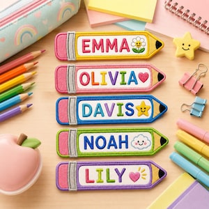 May include: Five embroidered pencil-shaped name tags with different names: Emma, Olivia, Davis, Noah, and Lily. Each tag features a different color scheme and a small embroidered icon, such as a flower or star. The tags are on a light wooden surface.