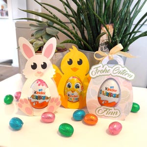 Peut inclure: Décorations de Pâques avec un lapin blanc, un poussin jaune et une boîte cadeau rose, chacun contenant un œuf Kinder Surprise. Des œufs en chocolat multicolores emballés dans du papier d'aluminium sont dispersés. Les mots "Frohe Ostern" et le nom "Finn" sont visibles.