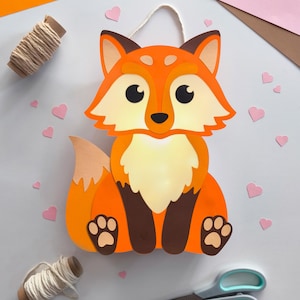 Op de afbeelding: Een handgemaakte oranje vossenversiering met een witte buik, bruine poten en een crèmekleurig ophangtouwtje. De vos is omgeven door roze hartvormige uitsparingen, oranje en roze papier en een spoel garen.
