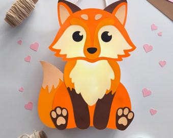 Könnte beinhalten: Handgefertigte orangefarbene Fuchskarte mit weißem Bauch, braunen Pfoten und einer cremefarbenen Aufhängeschnur. Der Fuchs ist von herzförmigen rosa Ausschnitten, orangefarbenem und rosa Papier und einer Garnrolle umgeben.