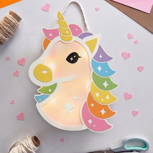 Könnte beinhalten: Ein Wandbehang in Form eines Einhornkopfes aus geschichtetem Papier mit integrierter Beleuchtung. Das Einhorn ist weiß mit einem gelben Horn, einer gelben Schnauze und einer bunten Mähne in Pink, Lila, Blau, Grün und Orange mit Sternausschnitten.