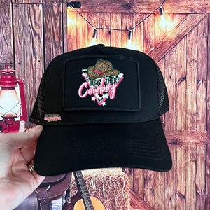 May include: Black trucker hat with a black and brown leopard print patch that says "Hey There Cowboy" in pink and green letters.