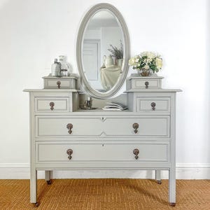May include: A light grey wooden vanity dresser featuring an oval mirror, two large drawers, and two smaller upper drawers on each side. The piece is accented with dark metal drop pulls and bead detailing along the drawer edges and mirror frame.