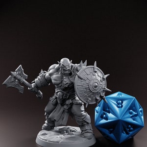 Peut inclure: Figurine d'orc gris avec une hache et un bouclier, à côté d'un grand dé D20 bleu. L'orc est musclé et détaillé, avec une expression féroce. Le bouclier a des pointes sur le bord. Le dé est d'un bleu vif.