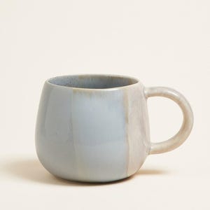 Op de afbeelding: Een keramische mok met een afgeronde body en een handvat. De mok is voornamelijk lichtblauw met een verticale beige streep. De binnenkant van de mok is blauw gespikkeld. De mok is ontworpen voor warme of koude dranken.