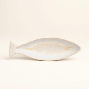 Könnte beinhalten: Eine keramische Fisch-Servierplatte. Die Platte ist weiß mit einem hellbeige Innenraum und einem flossenartigen Schwanz. Die Platte ist für die Aufnahme von Lebensmitteln konzipiert und eignet sich zum Servieren von Vorspeisen oder Snacks.