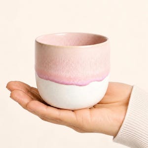Pode incluir: Uma pequena chávena de cerâmica arredondada com um design de dois tons. A parte superior é de um rosa suave e suave com um acabamento mate texturizado, enquanto a parte inferior é de um branco sólido e liso. As duas cores encontram-se numa linha.