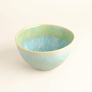Puede incluir: Un cuenco de cerámica con un esmalte azul claro y verde. El cuenco tiene una forma redondeada con un borde color crema. El interior del cuenco hace la transición de un azul claro a un tono verde.