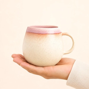 Könnte beinhalten: Eine gesprenkelte, cremefarbene Keramiktasse mit runder Form und kleinem Henkel. Der Rand der Tasse hat einen Farbverlauf von Rosa und Rot. Die Tasse wird in der Hand einer Person vor einem neutralen Hintergrund gehalten.