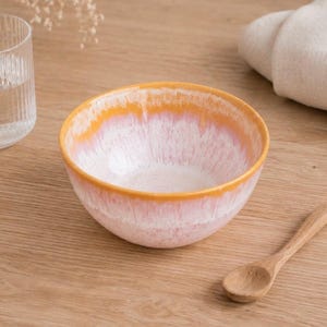 Könnte beinhalten: Eine Keramikschale mit weißem Innenraum und einem rosa-orangenen Farbverlauf am Rand. Ein Holzlöffel liegt auf einer Holzoberfläche neben der Schale. Ein Glas Wasser ist im Hintergrund zu sehen.