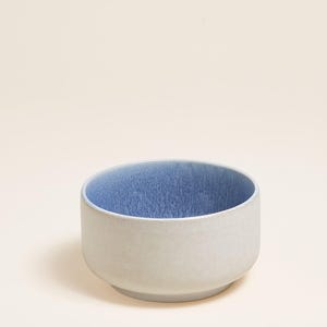 Pode incluir: Uma tigela de cerâmica com exterior cinza claro e interior azul. A tigela tem formato arredondado com uma borda ligeiramente alargada. O interior azul tem uma aparência texturizada.
