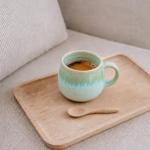 Può includere: Una tazza di ceramica turchese con caffè scuro, su un vassoio di legno con un piccolo cucchiaio di legno. La tazza ha un manico e una glassa verde chiaro. Il vassoio è rettangolare con bordi arrotondati.