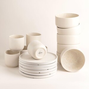 Può includere: Una collezione di stoviglie in ceramica bianco sporco, tra cui piatti, ciotole e tazze. I piatti sono impilati e le ciotole sono in una torre. Le tazze sono sparse intorno ai piatti. La parola "alma" è stampata sul fondo di una delle tazze.