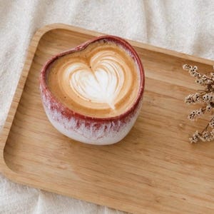 May include: A heart-shaped coffee cup with a latte design, featuring a heart shape in the foam. The cup is red and white, sitting on a wooden tray. A sprig of dried flowers is to the right.