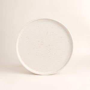 Könnte beinhalten: Ein runder, cremefarbener Keramikteller mit gesprenkelter Oberfläche. Der Teller hat einen leicht erhöhten Rand und ein mattes Finish. Die neutrale Farbe und das schlichte Design machen ihn für verschiedene Tischarrangements geeignet.