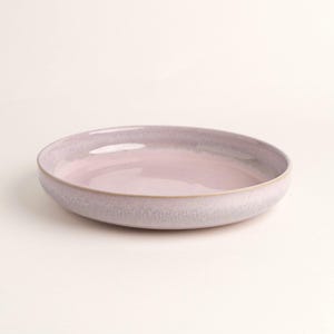 Pode incluir: Uma tigela redonda e rasa de cerâmica com um interior rosa suave e abafado e uma borda texturizada roxa clara. A tigela tem um design simples e elegante com uma borda sutil marrom dourado. O fundo é de cor branco sujo.