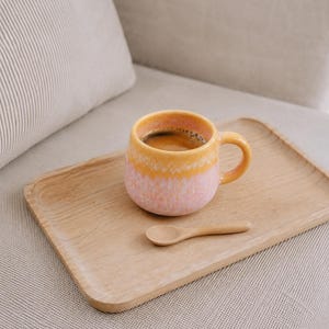 Könnte beinhalten: Ein Keramikbecher mit abgerundeter Form, der oben einen gelben Farbverlauf und an der Basis ein gesprenkeltes rosa Finish aufweist, gefüllt mit dunklem Kaffee. Der Becher steht auf einem rechteckigen Holztablett neben einem kleinen Holzlöffel.