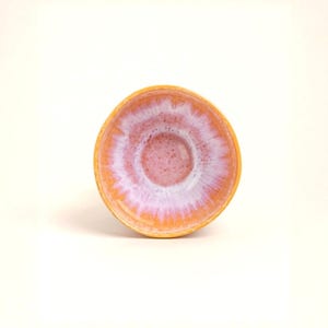 Può includere: Una piccola ciotola rotonda in ceramica con una vivace combinazione di colori. L'esterno è arancione caldo, mentre l'interno passa da un centro rosa tenue a un bordo bianco e lavanda. Il design della ciotola è semplice ed elegante.