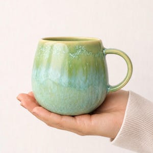 Könnte beinhalten: Eine Keramiktasse mit rundem Körper und Henkel. Die Tasse hat einen Farbverlauf aus Grün- und Blautönen mit glänzendem Finish. Die Tasse wird in der Hand einer Person vor weißem Hintergrund gehalten.