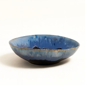 Op de afbeelding: Een handgemaakte keramische kom met een opvallend blauw glazuur. De kom heeft een donkerblauwe binnenkant die vervaagt naar een lichter blauw met een gouden rand. De buitenkant is donker, bijna zwart. Deze decoratieve kom is perfect om te serveren of tentoon te stellen.