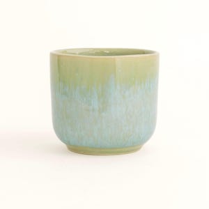 Può includere: Un bicchiere in ceramica con base arrotondata e lati dritti. La tazza ha una combinazione di colori sfumati, che passa da un verde chiaro nella parte superiore a un blu pallido nella parte inferiore. La tazza è progettata per le bevande.