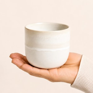 Könnte beinhalten: Eine kleine, runde Keramiktasse mit einer glatten, matten Oberfläche in Off-White- und Cremetönen. Das Gefäß hat ein schlichtes, henkelloses Design mit einem dezenten Farbverlauf, der von einer helleren Basis zu einem wärmeren Ton übergeht.