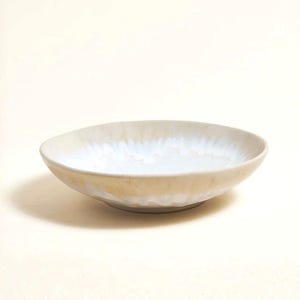 Könnte beinhalten: Eine cremefarbene Keramikschale mit perlweißem Innenraum. Die breite, flache Schale hat einen unregelmäßigen Rand und einen strukturierten Boden. Das Design ist minimalistisch und elegant. Die Schale eignet sich zum Servieren von Speisen oder als Dekorationsartikel. Durchmesser: ca. 20 cm.