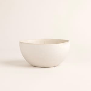 Puede incluir: Un cuenco de cerámica blanco roto con forma redondeada. El cuenco tiene una superficie lisa y mate y una amplia abertura. El color neutro y el diseño minimalista lo hacen adecuado para diversos usos, desde servir alimentos hasta guardar objetos decorativos.