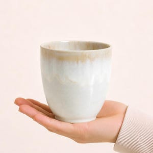 Könnte beinhalten: Ein Keramikbecher mit weißem Boden und hellbraunem Rand. Der Becher hat eine glatte, glänzende Oberfläche und wird in der Hand einer Person gehalten. Der Becher ist für Getränke konzipiert.