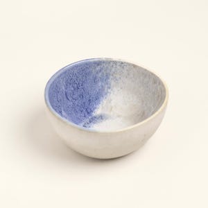 Könnte beinhalten: Eine kleine Keramikschale mit weißer Außenseite und blau-weißem Innenraum. Die blaue Farbe hat ein strukturiertes, gesprenkeltes Aussehen. Die Schale ist rund und steht auf einer weißen Oberfläche.