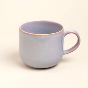 Può includere: Una tazza in ceramica con corpo arrotondato e manico curvo. L'esterno presenta una morbida sfumatura di toni lavanda e azzurro, mentre il bordo è accentuato da una sottile linea marrone dai toni caldi che aggiunge un contrasto delicato.