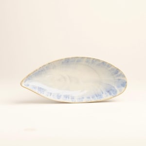 Peut inclure: Un plat en céramique en forme de feuille avec un glaçage bleu clair et blanc. Le plat a un bord légèrement surélevé et une finition lisse et brillante. La forme générale est allongée et organique.