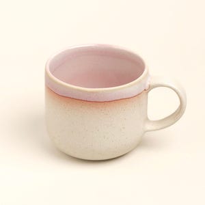 Può includere: Una tazza in ceramica con una base color crema maculata che sfuma in uno smalto rosa tenue sul bordo. La tazza presenta un corpo arrotondato, un interno liscio e un singolo manico curvo sul lato destro, caratterizzata da un design funzionale.