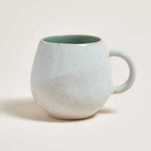 Peut inclure: Une tasse en céramique blanche avec un glaçage moucheté et un intérieur bleu clair. La tasse a un corps arrondi et une anse.