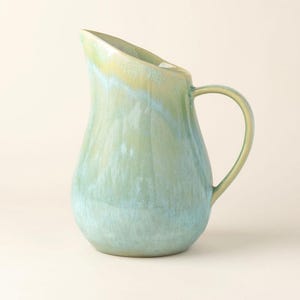 May include: A ceramic jug with a rounded body and a curved handle. The jug has a gradient of colours, transitioning from a light blue at the base to a pale green and yellow at the top. The jug is likely used for serving beverages.