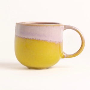 Könnte beinhalten: Eine Keramik-Tasse mit rundem Boden und großem Henkel. Die Tasse ist in zwei Bereiche unterteilt: Die untere Hälfte ist gelb und die obere Hälfte ist hellrosa. Die Tasse ist für Heißgetränke konzipiert.