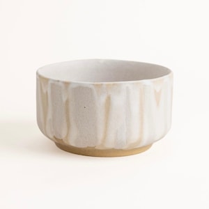 Pode incluir: Uma tigela de cerâmica redonda com uma base bege salpicada e um esmalte branco que escorre pelas laterais. A tigela apresenta uma abertura larga e uma base curta e robusta. A textura parece mate e terrosa, com variações visíveis no esmalte.