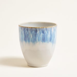 Könnte beinhalten: Ein Keramikbecher mit zweifarbigem Design. Die obere Hälfte des Bechers ist leuchtend blau und geht in eine hellere, fast weiße Farbe über. Der Rand hat einen dezenten braunen Akzent.
