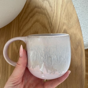 May include: A ceramic mug with a rounded shape and a curved handle. The exterior features a reactive glaze finish that transitions from a soft, pale pink at the base to a creamy, off-white color at the top, creating a subtle drip-like effect.