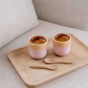 Pode incluir: Duas pequenas chávenas de café com gradiente rosa e amarelo cheias de café expresso estão numa bandeja de madeira, acompanhadas por duas pequenas colheres de madeira. As chávenas e colheres estão dispostas numa bandeja de madeira clara.