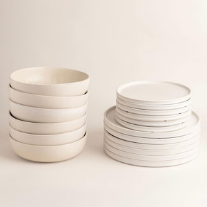 Könnte beinhalten: Ein Stapel weißer Keramikschalen und -teller. Die Schalen sind links und die Teller rechts gestapelt. Das Geschirr hat ein minimalistisches Design und ein mattes Finish.