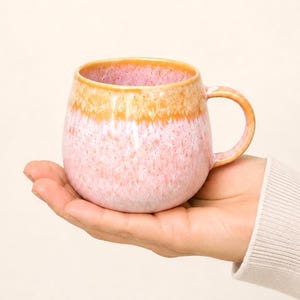 Könnte beinhalten: Eine rosa-orangefarbene Keramik-Tasse mit abgerundeter Form und Henkel. Die Tasse hat eine gesprenkelte Textur und einen Farbverlauf, der von Rosa zu Orange am Rand übergeht. Die Tasse wird in einer Hand gehalten.