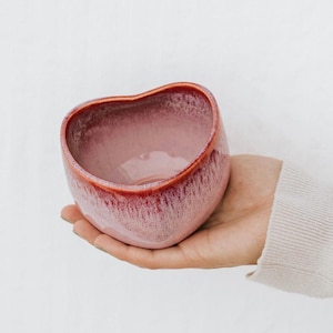 May include: A small, heart-shaped ceramic bowl with a reactive glaze finish. The bowl features a soft pink interior and exterior that transitions into a deep, reddish-brown rim, creating a distinct gradient effect across the curved surface.