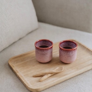 May include: Two small, pink and burgundy ceramic cups filled with a dark liquid sit on a light wooden tray. Two small wooden spoons are also on the tray. The tray is on a neutral-colored surface.