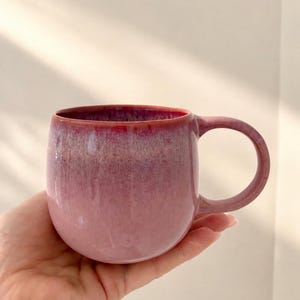 Könnte beinhalten: Ein abgerundeter Keramikbecher mit einer reaktiven Glasur, die von einem tiefen, gesättigten Magenta am Rand zu einem helleren, staubigen Altrosa an der Basis übergeht. Der Becher hat einen glatten, geschwungenen Körper und einen passenden Henkel.