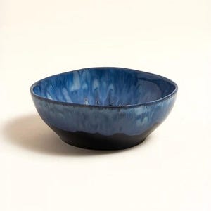 May include: A ceramic bowl with a distinctive design. The bowl has a dark blue exterior that fades into a lighter blue interior. The rim has an organic, slightly irregular shape, and the base is a solid black.