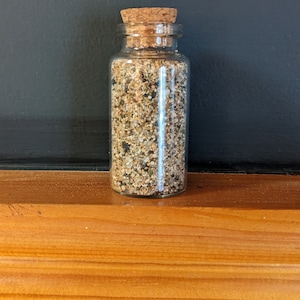 May include: A small glass jar with a cork lid filled with sand. The jar is sitting on a wooden surface.